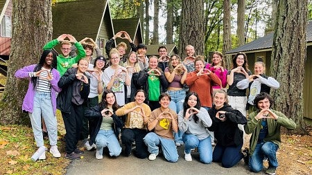 group of students posing among cabins in the woods and throwing the O