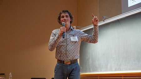 elliot nopp presenting at three minute thesis, gesturing with hand and holding mic